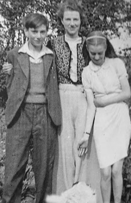 woman stands with arms around teenage boy and young girl, who reaches down to what looks like a dog.