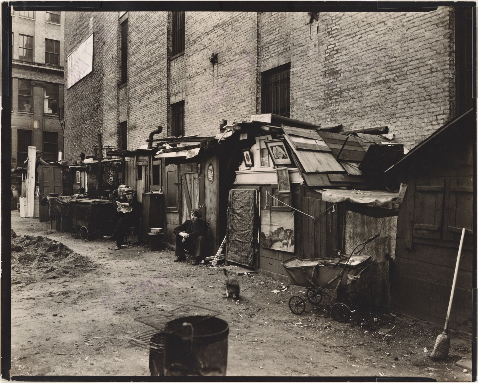 two men seated outside huts constructed against brick building.